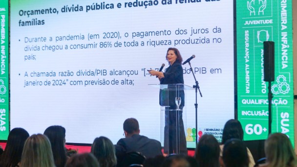 A imagem mostra uma mulher branca, de cabelos cacheados, vestida de azul. Ela está em cima de um palco, em frente a um púlpito, com um microfone na mão, falando para uma plateia. Atrás da mulher, um telão onde se leem diversas frases.